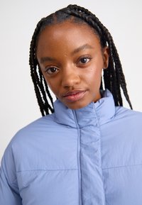 Veste rembourrée bleu clair avec un col haut, présentant une texture matelassée et des détails de couture à l'avant, offrant une apparence lisse et légère.