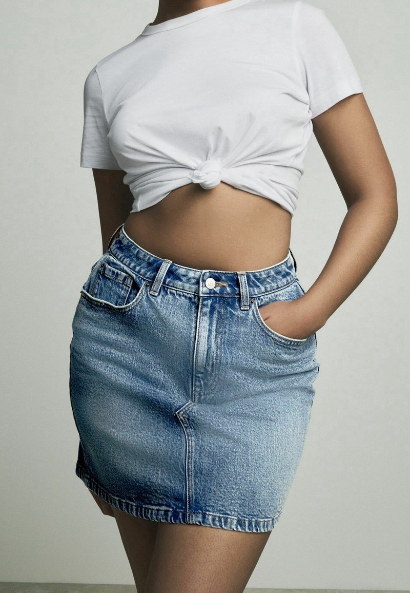 Verwaschener Jeans-Minirock in Hellblau mit Ausbleichungen, Fünf-Taschen-Design und Gürtelschlaufen. Kombiniert mit einem geknoteten weißen Baumwoll-T-Shirt.