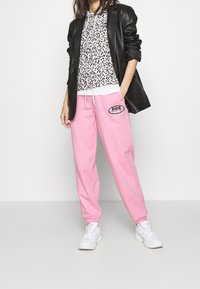 Person som bär rosa Puma-träningsbyxor, vita sneakers, en svart läderjacka och en vit hoodie med svarta leopardfläckar.
