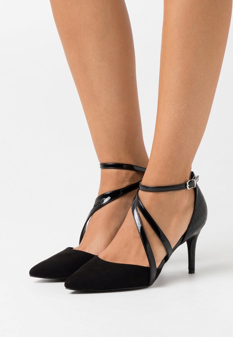 Chaussures à talons hauts noires avec un bout pointu, présentant une combinaison de lanières en suède et en cuir verni. Des brides de cheville croisées assurent un maintien.