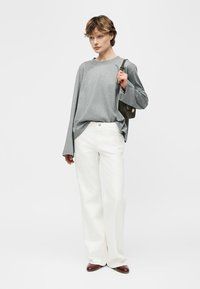 Personne portant une chemise grise ample à manches longues, un pantalon blanc à jambes larges, des chaussures marron, et portant un petit sac bandoulière vert foncé.