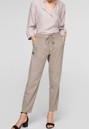 Vrouw draagt een licht mauve blouse met lange mouwen, ingestopt in beige broek met aantrekkoord en enkellengte, met zwarte open-toe blokhakken, staat met handen in de zakken.