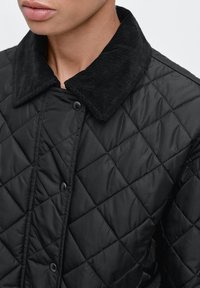 Zwarte doorgestikte jas met een corduroy kraag, drukknopen en een textuurrijke stofafwerking. Close-up toont het vakmanschap en ontwerp.