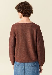 Brauner Strickpullover mit lockerer Passform, weitem Ausschnitt und strukturierten Ärmeln. Getragen mit einem Jeansrock.