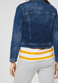 Denimjacka i mörkblått, kort design med sömdetaljer och knäppta ärmslut. Bärs över en vit och gulrandig skjorta.