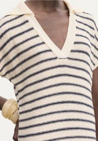 Personne portant un haut en tricot crème avec des rayures horizontales bleu marine et un col en V profond, accessoirisé avec des bracelets épais crème à un poignet.