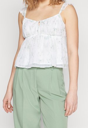 Mujer con blusa blanca sin mangas con patrones florales sutiles y pantalones verdes claros de talle alto y corte a medida.