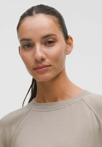 Pulover bej deschis cu guler rotund, mâneci raglan și textură moale. Subiectul are un ten natural cu pistrui, părul închis la culoare, dat pe spate.