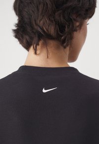 Personne aux cheveux courts et foncés portant un t-shirt noir avec un logo Nike blanc centré sur le haut du dos, sur un fond uni.