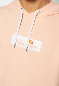 Sweat à capuche pêche clair avec cordons blancs et logo rectangulaire blanc avec un demi-cercle orange et rouge au-dessus d'un texte pâle.