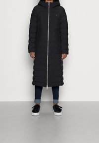 Abrigo acolchado negro con capucha, cremallera de largo completo y puños acanalados. Lleva puesto sobre unos vaqueros oscuros y zapatillas negras. Textura lisa e aislante.