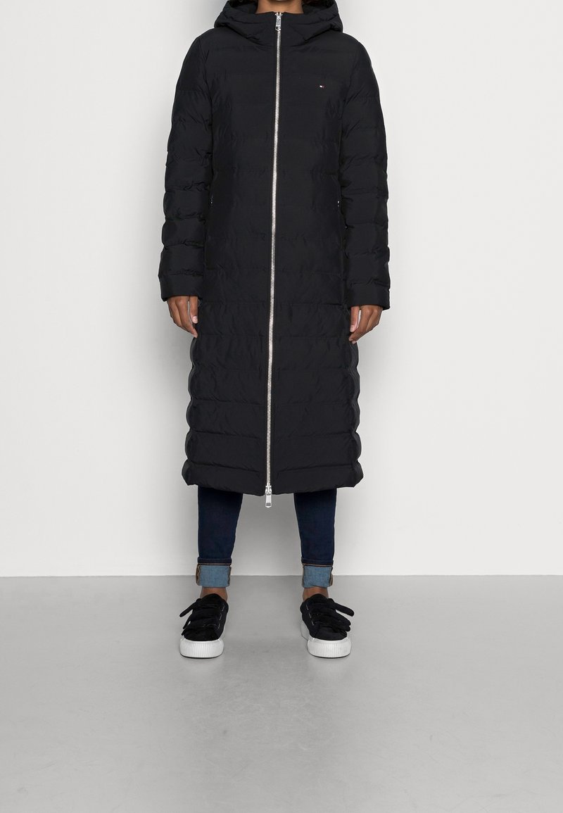 Abrigo acolchado negro con capucha, cremallera de largo completo y puños acanalados. Lleva puesto sobre unos vaqueros oscuros y zapatillas negras. Textura lisa e aislante.