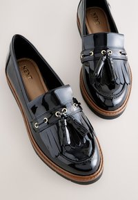 Mocassins en cuir verni noir avec un design à franges superposées, des accents en métal doré et des pompons décoratifs sur le devant, avec une semelle marron.