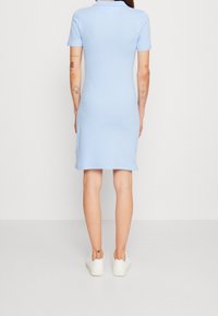 Lichtblauwe polojurk met korte mouwen, aansluitende pasvorm en geribbelde kraag. Draagt witte sneakers en heeft zichtbare tatoeages op de armen.