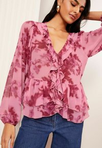 Rosa gemusterte Bluse aus leichtem Stoff, mit rüschigem V-Ausschnitt, langen Ärmeln und gestufter Detailgestaltung, kombiniert mit blauen Jeans.