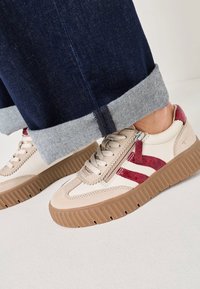 Baskets beiges avec des accents rouges, empeigne en cuir texturé et semelle en caoutchouc marron à nervures. Dotées d'une fermeture à lacets et d'un détail zippé.