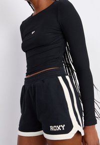 Top negro de manga larga acanalado combinado con un pantalón corto negro que presenta franjas laterales blancas y el logotipo "ROXY" en blanco en la parte inferior de la pierna.