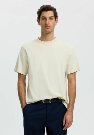 Jeune homme aux cheveux courts et bouclés portant un t-shirt beige uni et un pantalon bleu foncé avec une ceinture noire, debout avec une main dans la poche.