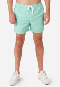 Vihreät uimalyhytshortsit, joissa on kiristysnauha vyötäröllä, käännetyt henkäykset ja sileä, kevyt kankaan rakenne. Yhdistetty valkoisiin lenkkareihin.