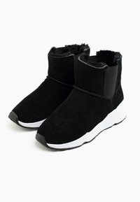 Botines de ante negros con una suela blanca, que cuentan con un interior acolchado y paneles laterales elásticos para facilitar su colocación. Textura suave, diseño de punta redonda.
