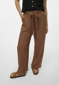 Pantalons larges marron avec un détail de ceinture à nouer, fabriqués à partir d'un tissu texturé. Associés à des sandales beiges, affichant un look confortable et décontracté.