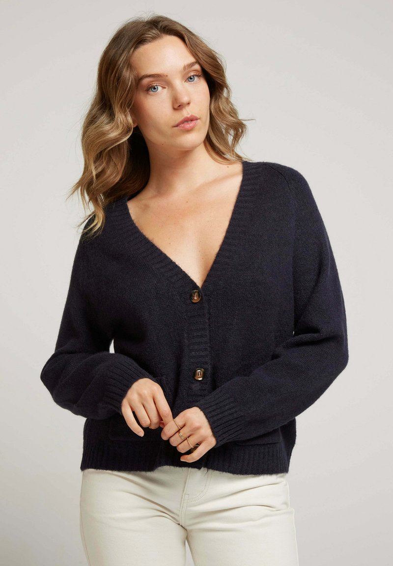 Gilet en tricot bleu marine avec un profond col en V et un ourlet côtelé, ornée de boutons en bois et de manches longues. Texture lisse, coupe décontractée.