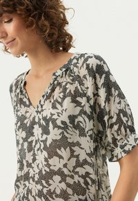 Let stadig, beige bluse med et sort blomsterdesign, V-udskæring og korte ærmer med rynkedetaljer. Glat tekstur.