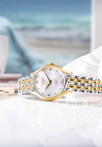 Reloj en tonos plateado y dorado con una esfera de nácar, adornada con seis diamantes, y una correa de eslabones que presenta colores metálicos alternos.
