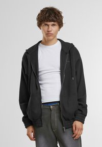 Schwarze Zip-Hoodie mit Kapuze zum Zuziehen, gerippten Bündchen und Fronttaschen. Getragen über einem weißen T-Shirt und kombiniert mit dunklen Hosen.