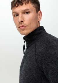 Veste en polaire zippée gris foncé avec un col haut, un tissu texturé et un tirette de fermeture éclair noire. Présente un design ajusté et des coutures discrètes.