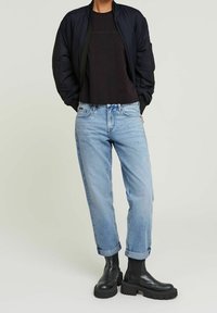 Černá bomber bunda, tmavé tričko, světlé modré džíny s ohrnutými manžetami a černé kotníkové boty s masivní podrážkou. Hladké materiály, volný střih.