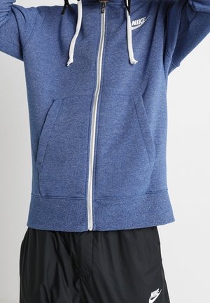 Personne portant un sweat à capuche zippé bleu Nike avec des cordons blancs et un short Nike noir, debout les bras levés.
