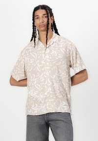 Kortermet, beige skjorte med knapper, dekorert med hvite abstrakte blomster mønstre og en glatt tekstur. Båret med grå jeans.