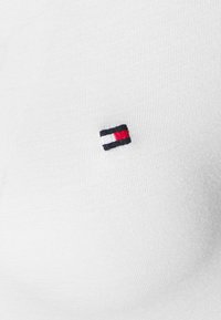 Camisa blanca de algodón con un pequeño logo de bandera bordado en negro, rojo y blanco en la parte izquierda del pecho. Textura suave.