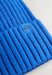 Berretto lavorato a maglia blu con coste verticali, caratterizzato da una superficie testurizzata e un piccolo patch con logo raffigurante un monte in bianco e nero.