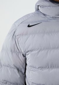 Lysegrå pufjakke med quiltede sektioner, der har et sort Nike-logo på skulderen. Materialet ser glat og let shiny ud.