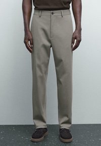 Pantaloni beige con taglio dritto, texture liscia e tasche frontali; abbinati a un top marrone scuro e scarpe scure.