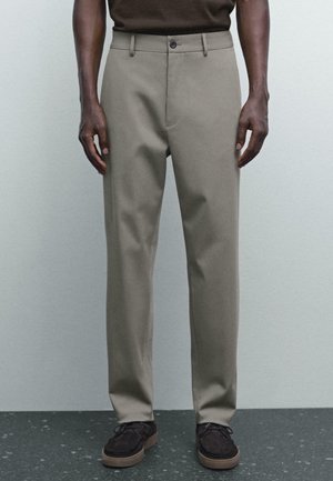 Pantalons beiges en tissu doux, présentant une coupe droite, des poches avant et une fermeture à un bouton. Portés avec des chaussures foncées.