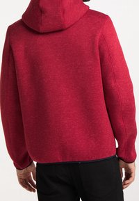 Punahuppari, joka on valmistettu teksturoidusta kankaasta. Siinä on huppu ja sivusaumat. Malli on rento, hieman väljällä istuvuudella ja resorireunuksilla hihoissa.