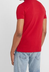 Czerwona koszulka polo z krótkim rękawem, wykonana z gładkiej bawełny, charakteryzująca się jednolitym kolorem, dopasowanym krojem i prostym kołnierzykiem. Założona z jasnoniebieskimi dżinsami.