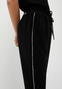 Czarne spodnie dresowe z elastycznym pasem, z bocznym paskiem z srebrnych cyrkonii dla dodatkowego efektu. Teksturowany materiał, luźny krój.