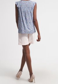 Blouse fleurie bleu clair avec manches volantées, associée à un short beige et des talons couleur nude. La blouse présente un détail froncé dans le dos.