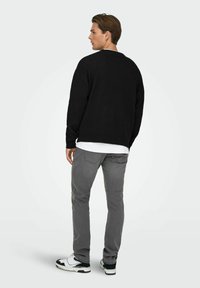 Pull en maille noire, jean gris et baskets noir et blanc. Le pull a une coupe décontractée et des poignets côtelés. Fond uni.