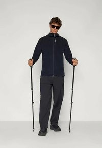 Zwarte fleece jas met hoge kraag, gecombineerd met donkere broek en zwarte wandelschoenen. De figuur houdt twee trekkingstokken vast.