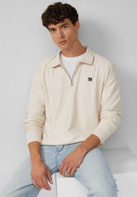 Cremfarbener Reißverschluss-Pullover mit gerippter Textur und Kragen. Mit einem Logo-Patch auf der Brust. Getragen über einem weißen T-Shirt und hellen Jeans.