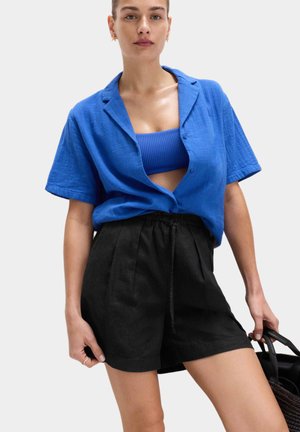 Frau trägt ein blaues Kurzarm-Hemdst mit Knöpfen über einem gerippten blauen Crop-Top und hoch taillierte schwarze Shorts, hält eine schwarze gewebte Tasche.