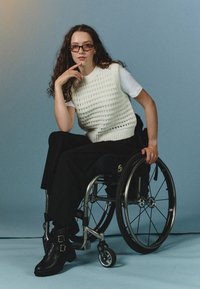 Un gilet bianco lavorato a maglia sopra una T-shirt bianca, pantaloni larghi neri e stivaletti neri alla caviglia. Carrozzina con telaio argentato e ruote nere. Sfondo azzurro chiaro.
