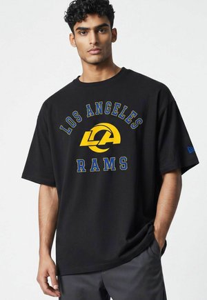 Jonge man met krullend donker haar draagt een zwart Los Angeles Rams T-shirt met geel en blauw teamlogo en tekst.