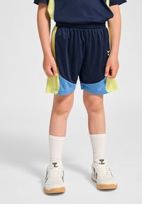 Marina atletiska shorts med gula och ljusblå accenter, elastisk midja och sidoventiler, ihop med vita strumpor och sneakers.