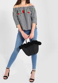Blusa off-the-shoulder nera e bianca a quadratini con ricami floreali rossi, abbinata a jeans skinny blu e una borsa nera con pompon.